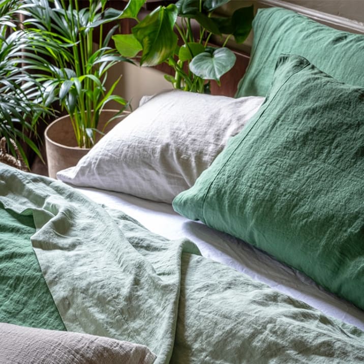 Green and white bedlinen on a bed in a bedroom with a parlour palm and monstera next to the bed